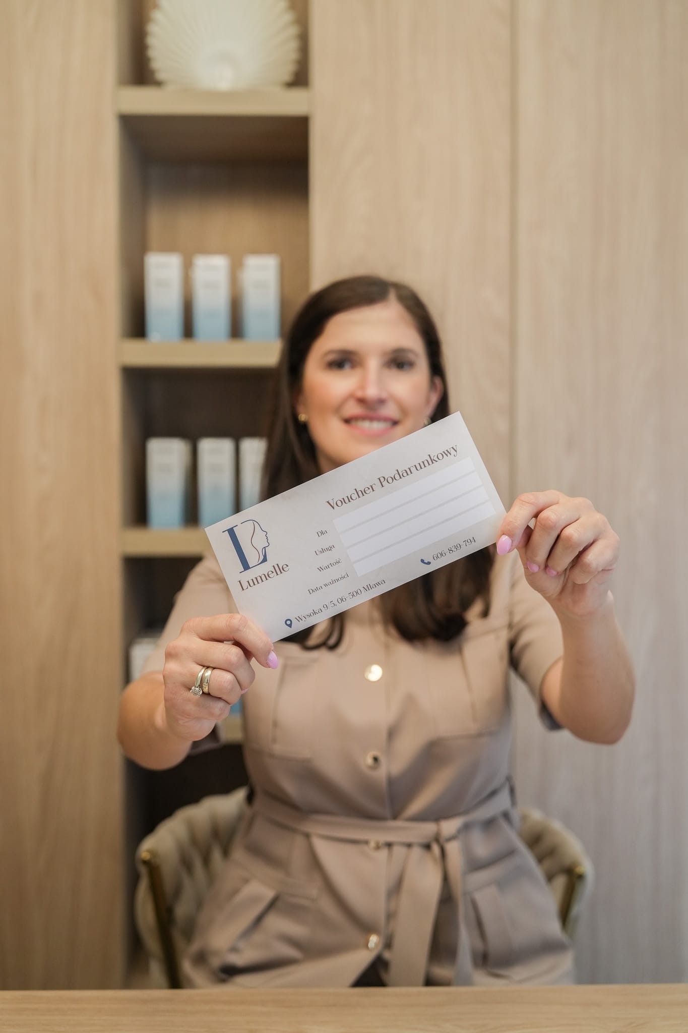 Specjalistka Lumelle pokazuje voucher podarunkowy gabinetu na tle drewnianych półek z kosmetykami