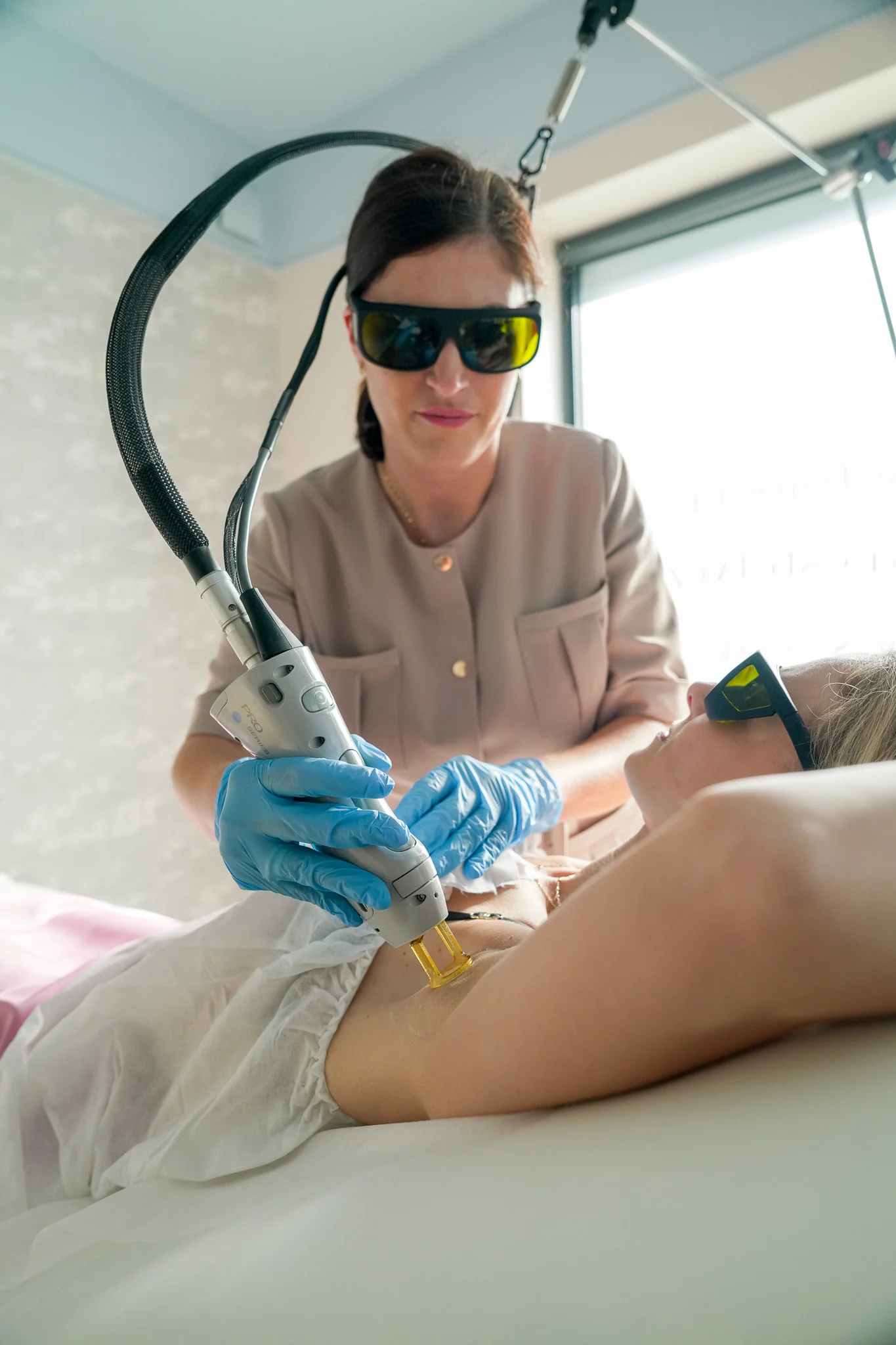 Specjalistka Lumelle w okularach ochronnych prowadzi głowicę lasera Candela w okolicy pachy klientki leżącej na łóżku zabiegowym; klientka też ma okulary ochronne