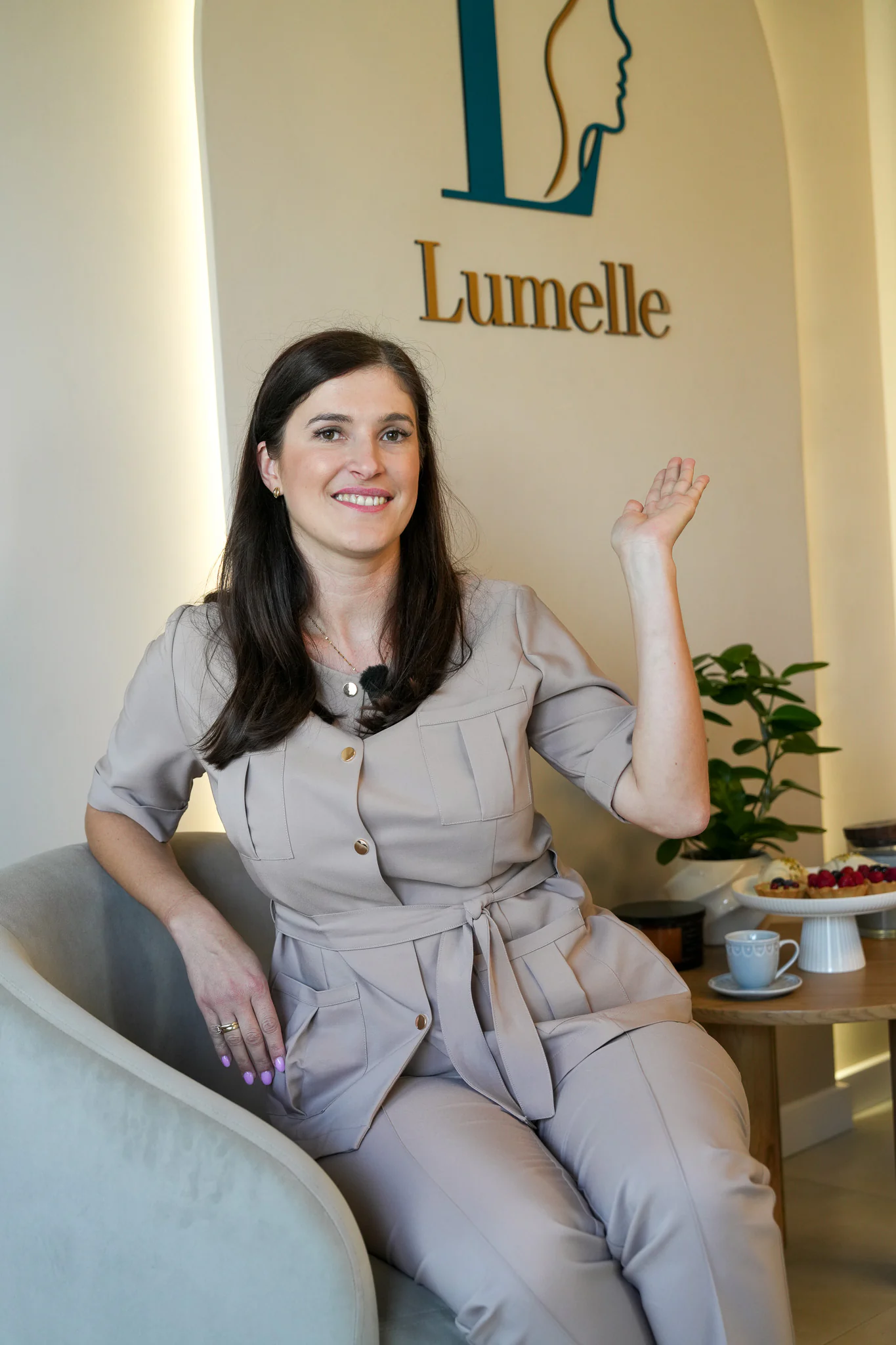 Specjalistka Lumelle w beżowym uniformie siedzi w fotelu w poczekalni gabinetu i unosi dłoń; za nią podświetlony łuk z logo Lumelle