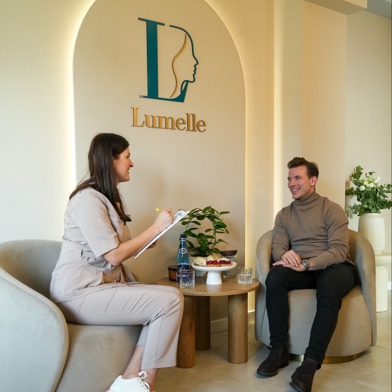 Konsultacja w Lumelle - specjalistka w beżowym uniformie rozmawia z klientem; oboje siedzą w fotelach pod podświetlonym łukiem z logo gabinetu