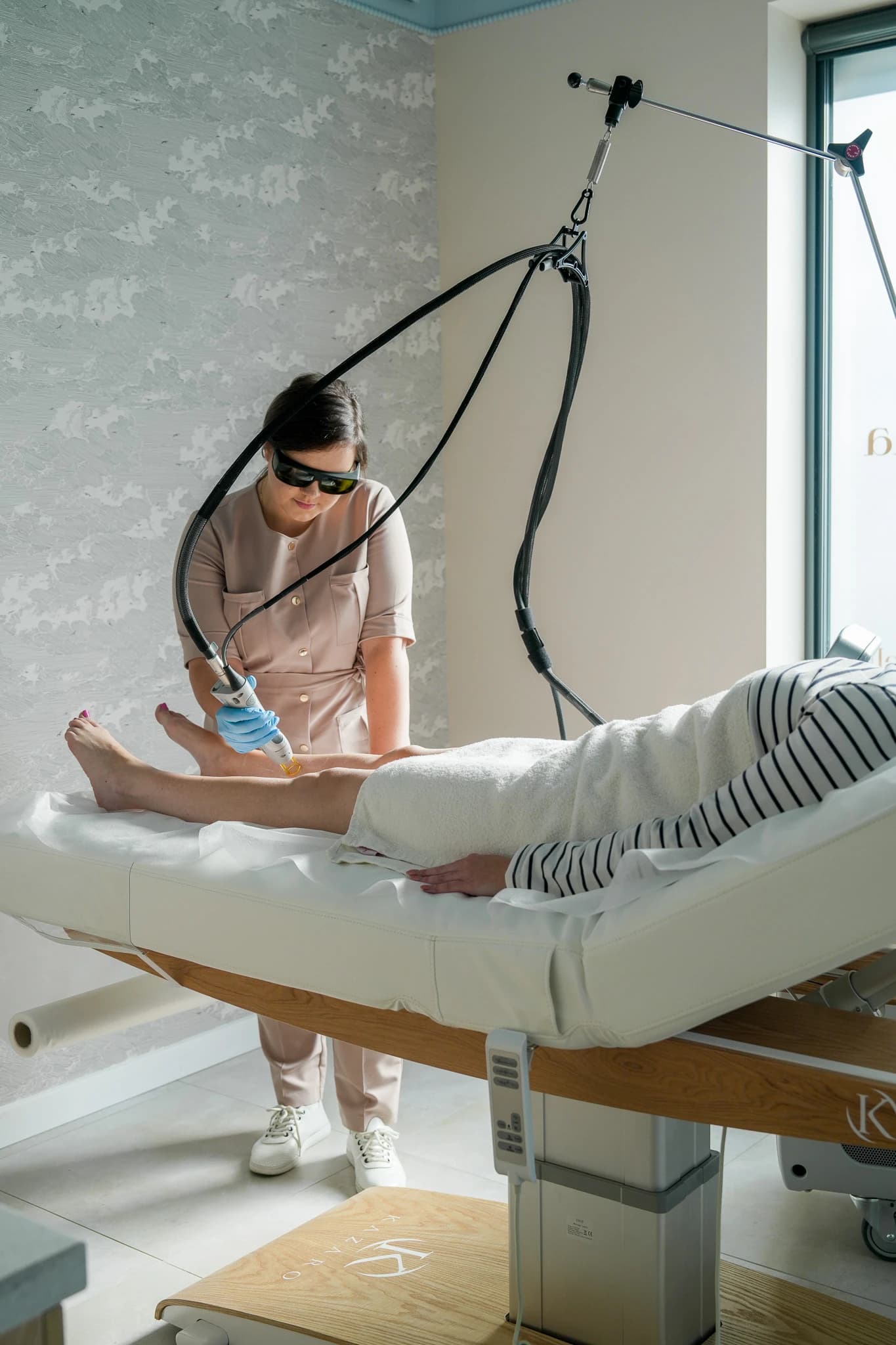 Depilacja laserowa Candela - specjalistka w okularach ochronnych przy łóżku zabiegowym prowadzi głowicę lasera po łydce klientki